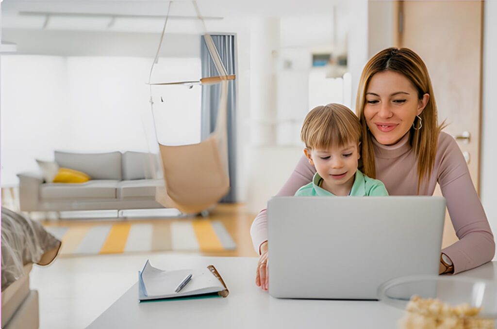 Happy mother managing work and family, typing on a computer while her child sits beside her at home.
